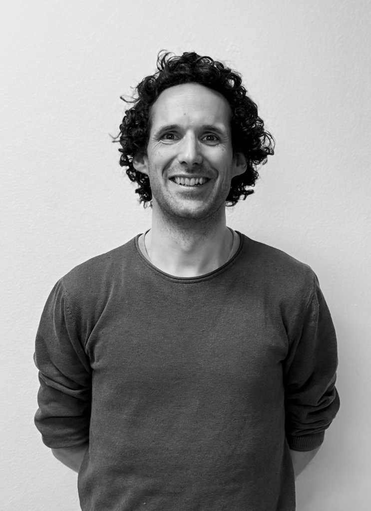 Portret van een man met krullend haar, glimlachend tegen een lichte achtergrond. Hij draagt een grijs T-shirt en steunt met zijn handen op zijn rug.